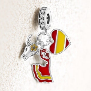 May include: A silver charm with a ring of clear stones. The charm features a bull's head, a heart with red and yellow stripes, and a flamenco dancer in a red dress with yellow accents. The charm is likely a souvenir or a piece of jewelry.