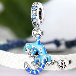 Könnte beinhalten: Ein silbernes Charm-Armband mit einem blauen und weißen Emaille-Gecko-Anhänger. Der Anhänger hat ein kleines rundes Schild mit der Aufschrift "Viel Glück".