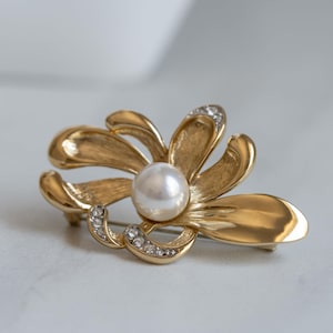 May include: A gold-toned flower brooch with a large, round, white pearl center. The brooch features curved petals and is accented with small, clear crystals. The brooch is a jewelry item.