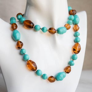May include: A turquoise and amber beaded necklace displayed on a white mannequin bust. The necklace features a mix of round and oblong beads in a two-strand design. The beads are strung on a gold-coloured chain.