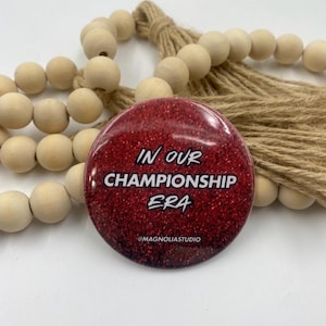 Puede incluir: Un botón redondo de purpurina roja con el texto blanco "IN OUR CHAMPIONSHIP ERA". El botón está rodeado de cuentas de madera y una cuerda de fibra natural. El texto "@MAGNOLIASTUDIO" está en la parte inferior.