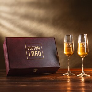 May include: A wooden box with a burgundy finish and a gold "CUSTOM LOGO" design, next to two champagne flutes filled with a golden liquid. The flutes also have a "CUSTOM LOGO" design. The items are on a wooden surface.