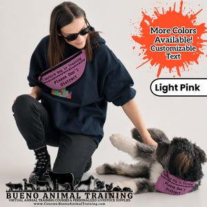May include: A light pink fanny pack featuring the black text Service Dog in Training Please Don't Distract. The fanny pack is worn across the chest by a person in a dark blue hoodie and black pants, while a dog wears a matching light pink bandana.