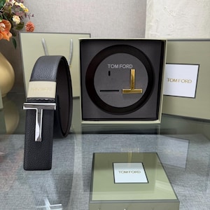 May include: A black leather belt with a silver-tone buckle and a gold-tone "T" logo. The belt is displayed with its packaging, which includes the "TOM FORD" logo. The belt is approximately 1.5 inches wide.
