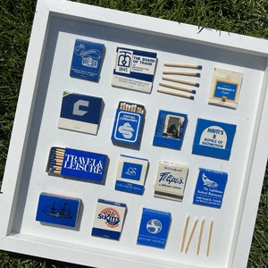 Può includere: Una collezione incorniciata di copertine di fiammiferi vintage, con disegni blu e bianchi con vari nomi di aziende e loghi, tra cui "Travel & Leisure", "The Board of Trade", "Haiti's 8 Hotels of Distinction" e "Royal St. Kitts Casino".