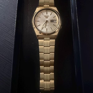 May include: A gold-toned Seiko automatic wristwatch with a matching gold band. The watch face is white with gold hands and markers, and the day and date are displayed. The watch is presented in a black box.