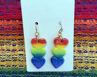 Könnte beinhalten: Ein Paar Regenbogen-Herz-Ohrringe. Jeder Ohrring hat drei Herzen, ein rotes, ein gelbes und grünes und ein blaues. Die Herzen sind glitzernd und haben eine Glitzer-Oberfläche.