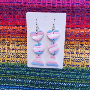 May include: A pair of dangle earrings with a transgender pride flag design. The earrings feature a heart, a circle, and a triangle, each with alternating stripes of blue, pink, and light blue.