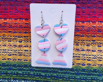 May include: A pair of dangle earrings with a transgender pride flag design. The earrings feature a heart, a circle, and a triangle, each with alternating stripes of blue, pink, and light blue.