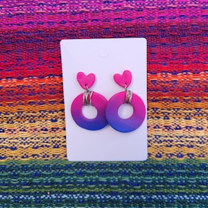 Può includere: Un paio di orecchini in gradiente di colore rosa e viola con un ciondolo a forma di cuore in cima e un anello di filo d'argento al centro.