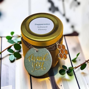 May include: A small glass jar of Peppermint Infused Honey with a gold-colored metal lid. A round, sage green tag with the gold-embossed text Thank you is tied to the jar with twine, alongside a small, gold-toned honeycomb-shaped metal charm.