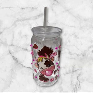 Pode incluir: Um copo de plástico transparente com um design de vaca marrom e branca e detalhes rosa. O copo tem um canudo e é decorado com contas rosa.