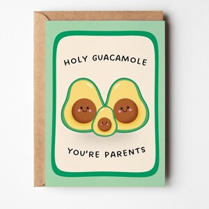 Op de afbeelding: Een wenskaart met een lichtgroene rand op een crèmekleurige achtergrond, met drie getekende avocado's met lachende gezichten. De tekst luidt HOLY GUACAMOLE YOU'RE PARENTS in zwarte hoofdletters. De kaart zit in een bruine kraftpapieren envelop.