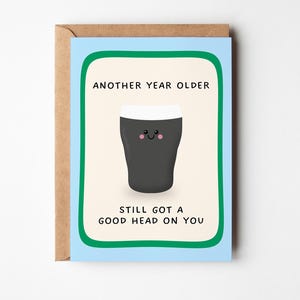 May include: A greeting card with a light blue border and a cream-coloured centre featuring a dark grey pint glass illustration with a white foam head and a smiling face. The text reads: ANOTHER YEAR OLDER, STILL GOT A GOOD HEAD ON YOU.