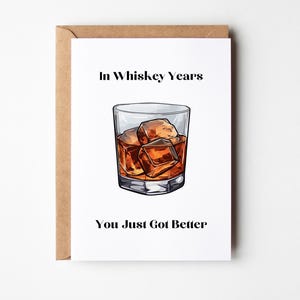 Könnte beinhalten: Eine weiße Grußkarte mit braunem Umschlag, die eine mittige Illustration eines Whiskeyglases mit Eiswürfeln zeigt. Der Text über dem Glas lautet In Whiskey Years und der Text unter dem Glas lautet You Just Got Better in schwarzer Schrift.