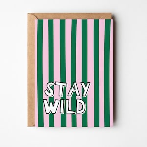 Könnte beinhalten: Eine rechteckige Grußkarte mit einem vertikalen Streifenmuster in abwechselnden Tönen von Waldgrün und Hellrosa. Der Text STAY WILD ist in fetten, weißen Großbuchstaben mit schwarzer Umrandung mittig auf der Vorderseite der Karte gedruckt.