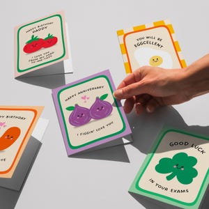 A hand is holding a greeting card with a colorful design featuring various fruits and vegetables, including tomatoes, eggplants, and a clover.