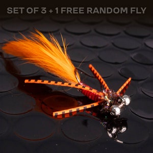 May include: A close-up of a fishing fly with orange and black details. The fly features a fluffy orange tail and a body with orange, black, and silver elements. The text "SET OF 3 + 1 FREE RANDOM FLY" is visible at the top.