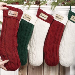 May include: Five cable-knit Christmas stockings in red, green, and white, each with a name tag. The stockings are displayed on a white mantelpiece decorated with festive greenery and pinecones, creating a cosy holiday scene.