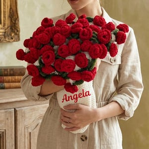 May include: A bouquet of red crochet roses in a white vase with the name "Angela" embroidered on it. The bouquet is held by a person wearing a beige linen shirt. The background includes a wooden cabinet, a mirror, and a window.