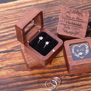 May include: Two wooden ring boxes with engraved details. One box has a clear lid and the other has a clear window. The box with the clear lid has two rings inside. The box with the clear window has the initials "R&G" engraved inside a heart.