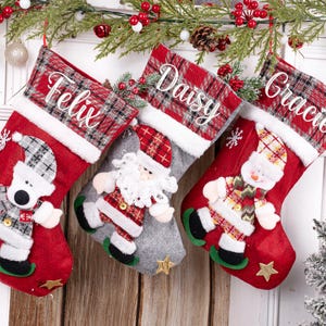 May include: Three Christmas stockings hang on a white door. The red stockings have white trim and are decorated with a bear, Santa, and a snowman. Each stocking has a name: Felix, Daisy, and Gracie.