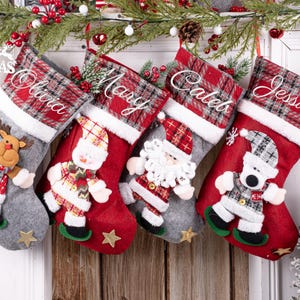 May include: Four Christmas stockings, each personalised with a name: Olivia, Mary, Caleb, and Jessica. Each stocking features a festive character: a reindeer, snowman, Santa Claus, and a dog. The stockings are red and grey with plaid detailing.