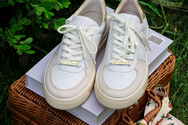 May include: Two white leather sneakers with gold accents. The left shoe has a gold tag that reads "Just Keep Running" and the right shoe has a gold tag that reads "Never give up!" The shoes are sitting on a white box with a brown wicker basket underneath.