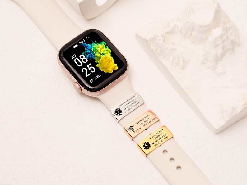 May include: A smartwatch with a cream strap displays the time and date. Three metal medical alert tags are attached to the strap. The watch face shows the time as 08:25 AM, with a colourful abstract background.