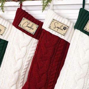 Puede incluir: Cuatro calcetines navide&ntilde;os de punto trenzado en verde, blanco y rojo. Cada calcet&iacute;n tiene una etiqueta de madera con un nombre: "Henry", "Charles", "Sophia" con una huella de pata y "Rachel".