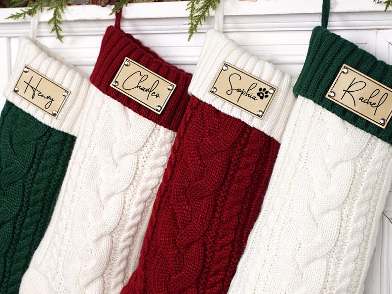 Puede incluir: Cuatro calcetines navide&ntilde;os de punto trenzado en verde, blanco y rojo. Cada calcet&iacute;n tiene una etiqueta de madera con un nombre: "Henry", "Charles", "Sophia" con una huella de pata y "Rachel".
