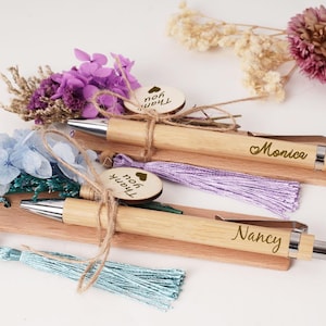 Könnte beinhalten: Zwei Holzstifte mit silbernen Akzenten, jeweils mit einem Namen und einem Herzsymbol graviert. Sie sind mit Quasten und kleinen Anhängern mit der Aufschrift "Thank you" verziert. Die Stifte sind mit Trockenblumen dekoriert.