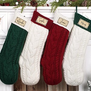 Puede incluir: Cuatro calcetines navide&ntilde;os de punto trenzado en verde, blanco y rojo cuelgan de una repisa blanca. Cada calcet&iacute;n tiene una etiqueta de madera con el nombre: Amelia, Sophia, Jason y Henry. Decoraci&oacute;n festiva.
