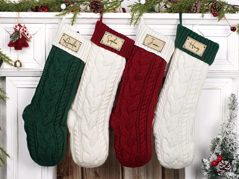 Puede incluir: Cuatro calcetines navide&ntilde;os de punto trenzado en verde, blanco y rojo cuelgan de una repisa blanca. Cada calcet&iacute;n tiene una etiqueta de madera con el nombre: Amelia, Sophia, Jason y Henry. Decoraci&oacute;n festiva.