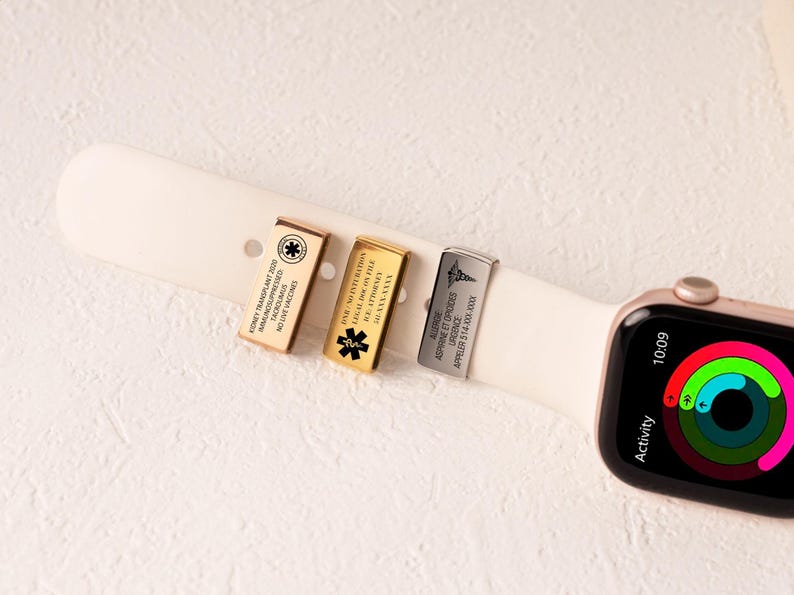 May include: Three metal medical alert tags attached to a white watch strap. The tags are gold, silver, and bronze, each with engraved medical information and symbols. The watch face displays the time and activity rings.