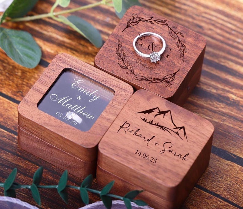 May include: Three wooden ring boxes with engraved designs. The first box has a floral wreath with the initials "L & G" and the date "21.MAY.2023". The second box has a window and the text "Emily & Matthew 08.07.23". The third box has a mountain range design and the text "Richard + Sarah 14.06.25".