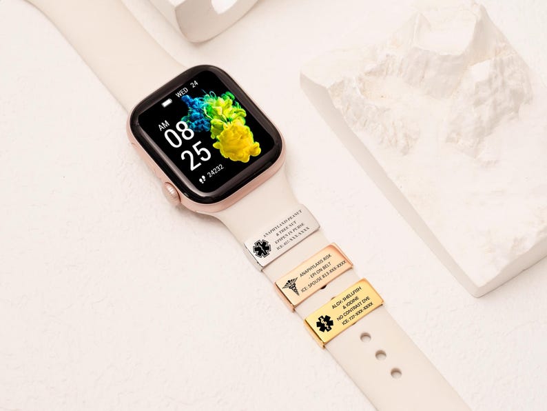 May include: A smartwatch with a cream-colored band displays the time and date. Three metal medical alert tags are attached to the band. The watch face shows the time as 8:25 AM, with a colorful abstract background.