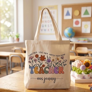 May include: A beige canvas tote bag with a floral design and the word "TEACHER" in colorful retro lettering. The text "Teach them, love them, watch them grow" is above the word "TEACHER", and "ms. jenny" is written below.