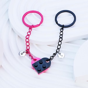 May include: A pink and black heart-shaped keyring with a black chain and a pink chain. The keyring has two small charms with the letters "G" and "L" on them.