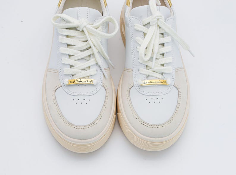 May include: A pair of white leather sneakers with gold metal accents. The left shoe has a gold tag that reads "Helman" and the right shoe has a gold tag that reads "Run with your heart".