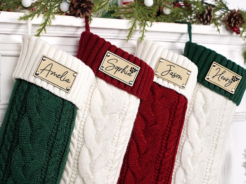 Puede incluir: Cuatro calcetines navide&ntilde;os personalizados en punto trenzado verde, rojo y blanco. Cada calcet&iacute;n tiene una etiqueta de madera con los nombres: Amelia, Sophia, Jason y Henry. La etiqueta de Sophia incluye una huella de pata.