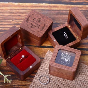 May include: Three wooden ring boxes with different designs. The first box has a lid with a floral design and the initials "E" and "M". The second box has a clear lid and a floral design with the names "Charlie" and "Sophia" and the date "Jun 24, 2024". The third box has a red velvet interior.