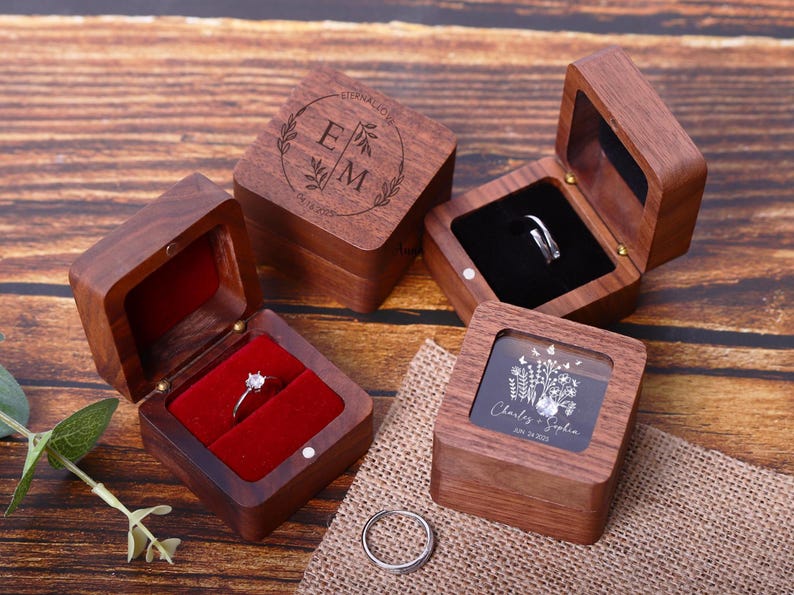 May include: Three wooden ring boxes with different designs. The first box has a lid with a floral design and the initials "E" and "M". The second box has a clear lid and a floral design with the names "Charlie" and "Sophia" and the date "Jun 24, 2024". The third box has a red velvet interior.