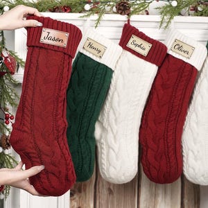 Puede incluir: Una colecci&oacute;n de cinco calcetines navide&ntilde;os de punto trenzado en rojo, verde y blanco, cada uno con una etiqueta con nombre personalizado. Los calcetines se exhiben en una repisa blanca con vegetaci&oacute;n festiva y pi&ntilde;as.