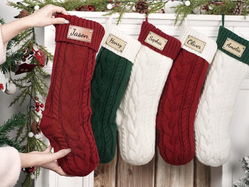 Puede incluir: Una colecci&oacute;n de cinco calcetines navide&ntilde;os de punto trenzado en rojo, verde y blanco, cada uno con una etiqueta con nombre personalizado. Los calcetines se exhiben en una repisa blanca con vegetaci&oacute;n festiva y pi&ntilde;as.