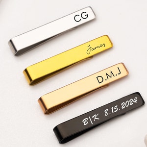 Può includere: Quattro fermagli per cravatta con diverse incisioni. Il fermaglio superiore è argentato e inciso con "CG". Il secondo fermaglio è dorato e inciso con "James". Il terzo fermaglio è in oro rosa e inciso con "D.M.J". Il fermaglio inferiore è nero e inciso con "B|K 8.15.2024".