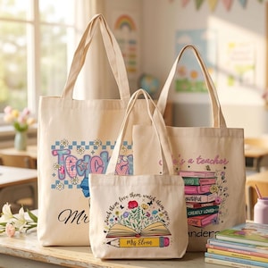 May include: Three beige canvas tote bags with teacher-themed designs. The largest bag has the word "Teacher" in a colorful floral design. The middle bag has an open book with flowers and the text "teach them love them, watch them grow". The smallest bag has a stack of books.