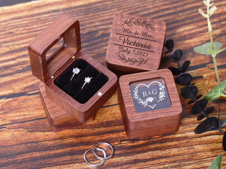 May include: Two wooden ring boxes with engraved details. One box has a clear lid and the other has a clear window. The box with the clear lid has two rings inside. The box with the clear window has the initials "R&G" engraved inside a heart.