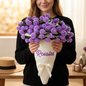 May include: A handmade bouquet of purple crochet roses in a white cone-shaped holder with a bow. The word "Rosales" is embroidered on the holder. The person is wearing a black sweater.