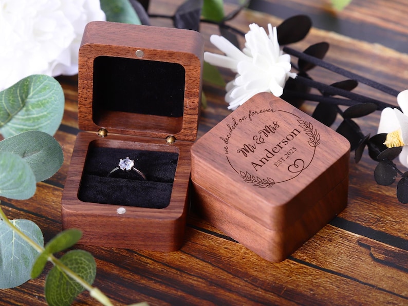 May include: A wooden ring box with a personalized inscription that reads "We decided on forever Mr. & Mrs. Anderson Est. 2023". The box is open and a ring with a single diamond is inside.